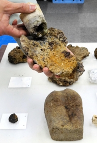 古墳時代中期ー後期の建物跡で見つかった鉄生産の名残。手に持っているのはふいごから風を送り込んだ土製の管「羽口」と精錬時に生じる不純物の鉄滓(てっさい)。手前右は砥石(といし)=津門大塚町遺跡 古墳時代中期ー後期の建物跡で見つかった鉄生産の名残。手に持っているのはふいごから風を送り込んだ土製の管「羽口」と精錬時に生じる不純物の鉄滓(てっさい)。手前右は砥石(といし)=津門大塚町遺跡