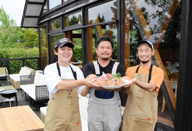オープンした店のテラスで、県内の農家が育てた野菜や果物を掲げる芦田賢太郎さん(中央)と共同経営者の森田航大さん(左)、加藤泰輔さん=神戸市西区神出町小束野