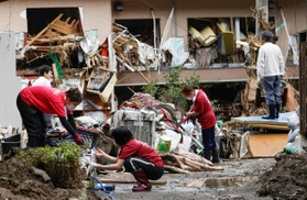 豪雨で氾濫した球磨川の水が押し寄せ、2階の窓から椅子や木材が飛び出した建物と、水に漬かった家財を片付ける家族ら=10日午前9時25分、熊本県八代市 豪雨で氾濫した球磨川の水が押し寄せ、2階の窓から椅子や木材が飛び出した建物と、水に漬かった家財を片付ける家族ら=10日午前9時25分、熊本県八代市