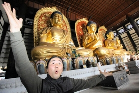 世界最大級の木造三大仏「但馬大仏」の前で仰天ポーズを取る阿瀬大典さん=香美町村岡区川会 世界最大級の木造三大仏「但馬大仏」の前で仰天ポーズを取る阿瀬大典さん=香美町村岡区川会