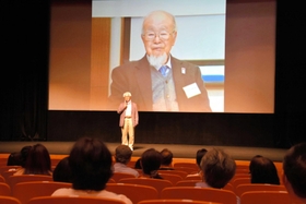 河合雅雄さんの写真を背景に、舞台であいさつする映画「森の学校」の西垣吉春監督=田園交響ホール 河合雅雄さんの写真を背景に、舞台であいさつする映画「森の学校」の西垣吉春監督=田園交響ホール