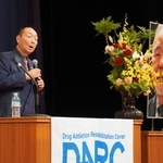 近藤恒夫さんの遺影が飾られた会場であいさつする、茨城ダルク代表の岩井喜代仁さん=27日午後、東京都台東区