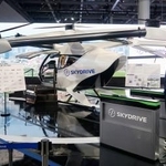スカイドライブがジャパンモビリティショーで展示する、空飛ぶクルマの模型と自動改札機。右は福沢知浩CEO=29日午前、東京都江東区の東京ビッグサイト