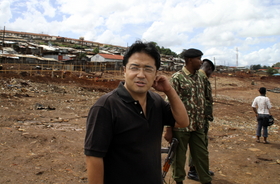 バラックが強制撤去され、更地になった場所に立つ宮田久也。後ろの武装警察官が自動小銃を手に警護にあたる=ケニア・ナイロビのキベラ・スラム バラックが強制撤去され、更地になった場所に立つ宮田久也。後ろの武装警察官が自動小銃を手に警護にあたる=ケニア・ナイロビのキベラ・スラム