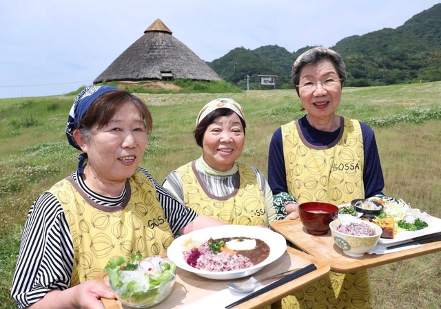 五斗長垣内遺跡の復元建物を背景に、自慢のメニューを手にする「まるごキッチン」の女性たち=淡路市黒谷 五斗長垣内遺跡の復元建物を背景に、自慢のメニューを手にする「まるごキッチン」の女性たち=淡路市黒谷