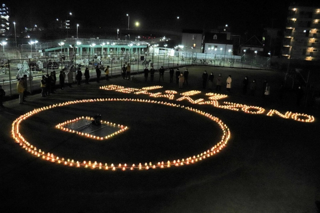 ろうそくの火で浮かび上がった「１・１７　ＢＥ　ＨＡＮＡＺＯＮＯ」を囲み、鎮魂の願いをささげる地域住民ら＝明石市西明石南町１
