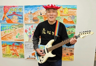 リニューアルした紙芝居をバックにギターを持つ福石健一さん＝明石市内