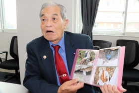 パラオで遺骨収集に参加した時の写真を手に、父の戦死などについて話す谷忠義さん＝淡路市役所
