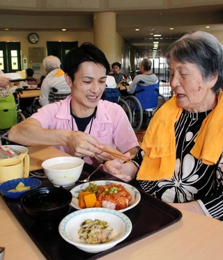 昼食時に入所者の女性に箸を手渡す湯本さん。「男前やし、話も上手。満点や」と女性＝神戸市西区櫨谷町長谷、「大慈弥勒園」