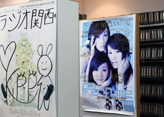２００７年発表のシングル「ポリリズム」の直筆サイン入りポスター（中央）。左は同じ中田ヤスタカ氏を音楽プロデューサーとするきゃりーぱみゅぱみゅのポスター＝神戸市中央区東川崎町１、ラジオ関西（撮影・長嶺麻子）