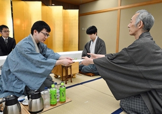 立会人の中田功八段（右）に封じ手を渡す永瀬拓矢九段（手前左）。中央は藤井聡太王位＝１９日午後、福岡県宗像市、宗像ユリックス