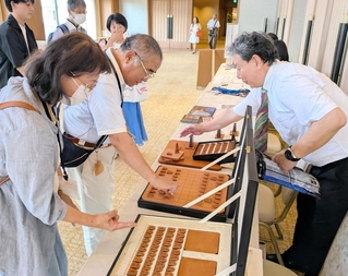 王位戦前夜祭に展示された牛革製の将棋盤と駒について説明する服部清隆さん（右）＝有馬グランドホテル