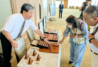 王位戦前夜祭に展示された牛革製の将棋盤と駒について説明する服部清隆さん（左）＝有馬グランドホテル