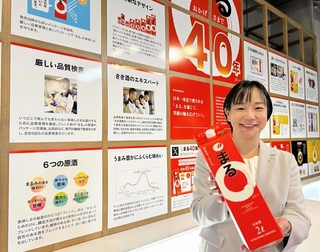「まる」の逸話を紹介するパネルが並ぶ会場で商品を持つ白鶴酒造の金子奈都子さん＝神戸市東灘区住吉南町４、同社資料館