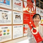 「まる」の逸話を紹介するパネルが並ぶ会場で商品を持つ白鶴酒造の金子奈都子さん＝神戸市東灘区住吉南町４、同社資料館
