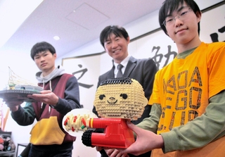 玩具のレゴでできた「ケンミン坊や」。灘中学・高校で、生徒たちから高村祐輝社長（中央）に渡された＝神戸市東灘区魚崎北町８