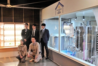 白鶴酒造の小規模醸造所の横にスパイスキューブ社の植物栽培装置（左）を設置し、記念撮影に応じる両社の関係者ら＝神戸市東灘区住吉南町４、白鶴酒造資料館館