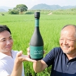 原料の酒米・山田錦の田を背に、日本酒「然土」を手にする藤原久和さん（右）と宝酒造の新宅恒基さん＝西脇市蒲江