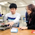 インプルーヴ代表の鈴木弘美さん（右）に見守られながら、専用の機械でリンキングを行う生徒＝神戸市灘区岩屋北町６、青陽灘高等支援学校