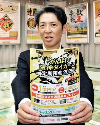 タイガース定期預金の過去のポスターを背に、今年の募集チラシを持つ尼崎信用金庫の潮成之介執行役員＝尼崎市西本町北通３
