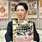 タイガース定期預金の過去のポスターを背に、今年の募集チラシを持つ尼崎信用金庫の潮成之介執行役員＝尼崎市西本町北通３