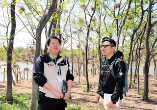 伊丹市みどり自然課の職員から話を聞く松永Ｋ三蔵さん（右）。頭上にはカワウが巣を作っている＝伊丹市昆陽池３