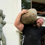 魚吹八幡神社に残る力石に挑戦したマーティンス・リシス選手。背後で三ノ宮卯之介の石像が見守る（大阪チキンレッグス提供）