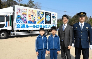 交通安全ポスターを印刷したトラック。（左から）ポスターを描いた岡井凰汰君と三宅杏汰君、平田優宇社長、大戸英治署長＝九会小学校