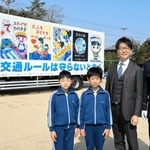 交通安全ポスターを印刷したトラック。（左から）ポスターを描いた岡井凰汰君と三宅杏汰君、平田優宇社長、大戸英治署長＝九会小学校