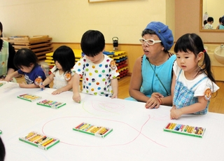 播州織の生地にクレヨンで絵を描く園児とアユミアダチさん（右から２人目）＝西脇こども園