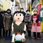 「みのる坊」人形を囲んで笑顔をみせる大日六商店街の店主ら＝神戸市中央区大日通６
