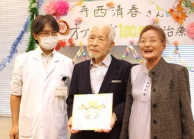 安井久晃医師（左）から色紙を受け取った寺西清春さんと妻の久美子さん＝神戸市立医療センター中央市民病院