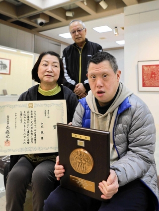 母和美さん（左）と藤原常貴さん（奥）に見守られて県知事賞の盾を手にする中尾徹さん＝三木市立堀光美術館