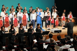 丘みどりさん（舞台中央）とともに、宍粟市歌「ここがふるさと」を披露する吹奏楽団と合唱団の団員ら＝山崎文化会館