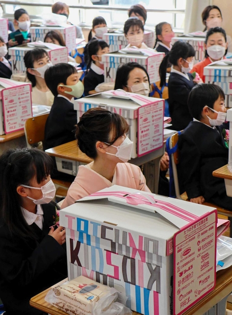 市から贈られた学用品の箱の前に座る新１年生と付き添いの保護者＝龍野小学校