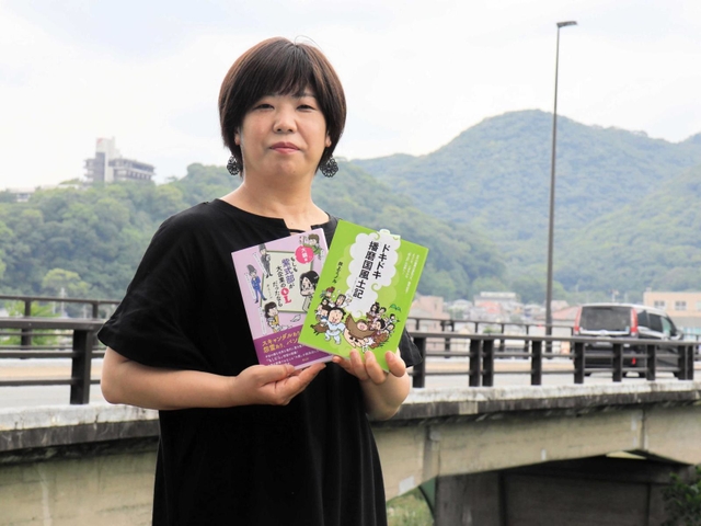 揖保川の流れを背に、著書をＰＲする井上ミノルさん。背後には白鷺山（左）と台山がある＝たつの市龍野町富永