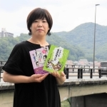 揖保川の流れを背に、著書をＰＲする井上ミノルさん。背後には白鷺山（左）と台山がある＝たつの市龍野町富永