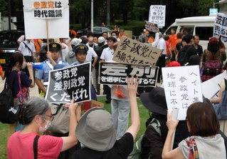 オレンジの服を着た参政党の陣営関係者に対し、プラカードを掲げて抗議する人たち。やじを飛ばす人もおり、双方の間にトラブルを警戒する警察官が立っていた＝17日午後、神戸市中央区加納町６、東遊園地