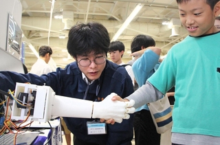 握手を握り返す装置に驚く来場者（右）と仕組みを説明する学生（中央）＝神戸市中央区浜辺通５、神戸サンボーホール
