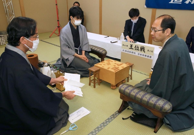 王位戦第２局で、立会人の深浦康市九段（左）から封じ手を示される木村一基王位（右）。左から２人目は藤井聡太七段＝１４日午前、札幌市、ホテルエミシア札幌