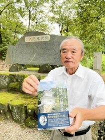 龍野公園の顕彰碑を背に、田中静壱の評伝小説を持つ筆者の横家伸一さん＝たつの市龍野町下霞城