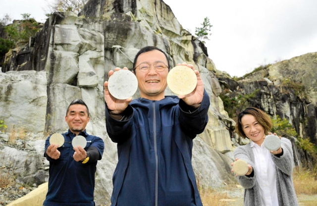 竜山石の採石場を背に返礼品のコースターをアピールする（左から）松下尚平さん、八木健吾さん、上谷奈穂子さん＝高砂市阿弥陀町魚橋