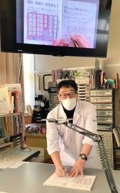 タブレットをアームで固定し、手元のプリントを映してオンライン授業の方法を説明する国眼教諭。教室にいる児童は大型画面を見て学ぶこともできる＝朝来市立竹田小（国眼教諭提供）