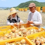 畑を背にタマネギの育ち具合を確かめる北川忠彦さん（右）と妻の由美子さん＝南あわじ市賀集福井