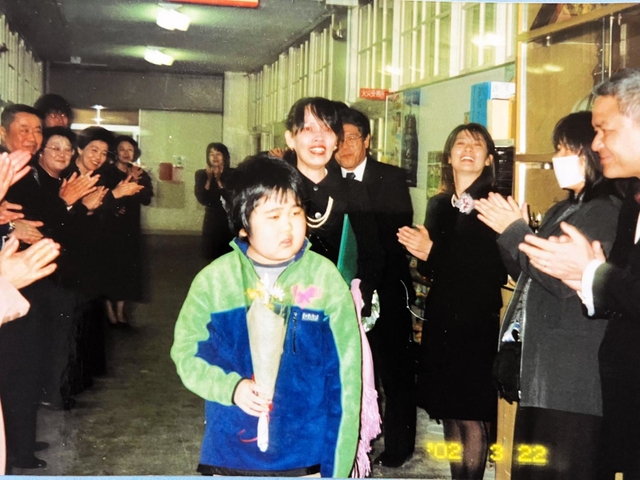 重病を乗り越え、鶴甲小学校の卒業式に臨む市川裕太さんと母玲子さん。先生や職員が祝福する＝２００２年、神戸市灘区（市川さん提供）