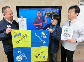 南海トラフ巨大地震の啓発動画を背に、災害時に使えるバンダナなどを手にする（左から）岩本吉正さん、小野多恵子さん、生駒良友さん＝神戸市灘区岸地通１