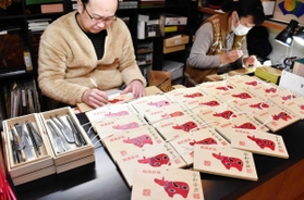 麦わら細工で「赤べこ」をあしらった絵馬を制作する神谷俊彰さん（左）＝かみや民芸店