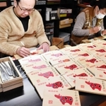 麦わら細工で「赤べこ」をあしらった絵馬を制作する神谷俊彰さん（左）＝かみや民芸店