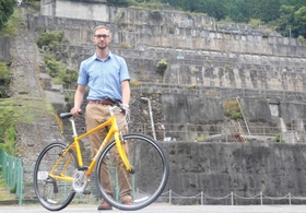 自転車を支えながら、神子畑選鉱場跡の前に立つケビンさん＝朝来市佐嚢