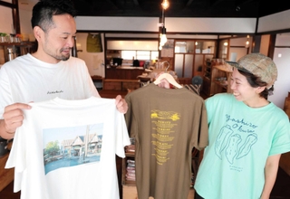 豊岡市日高町の地名や八代オクラがデザインされたＴシャツ。「農家さんがよく着ているオリーブ色は外せなかった」と話す冨樫実和さん（右）＝豊岡市日高町